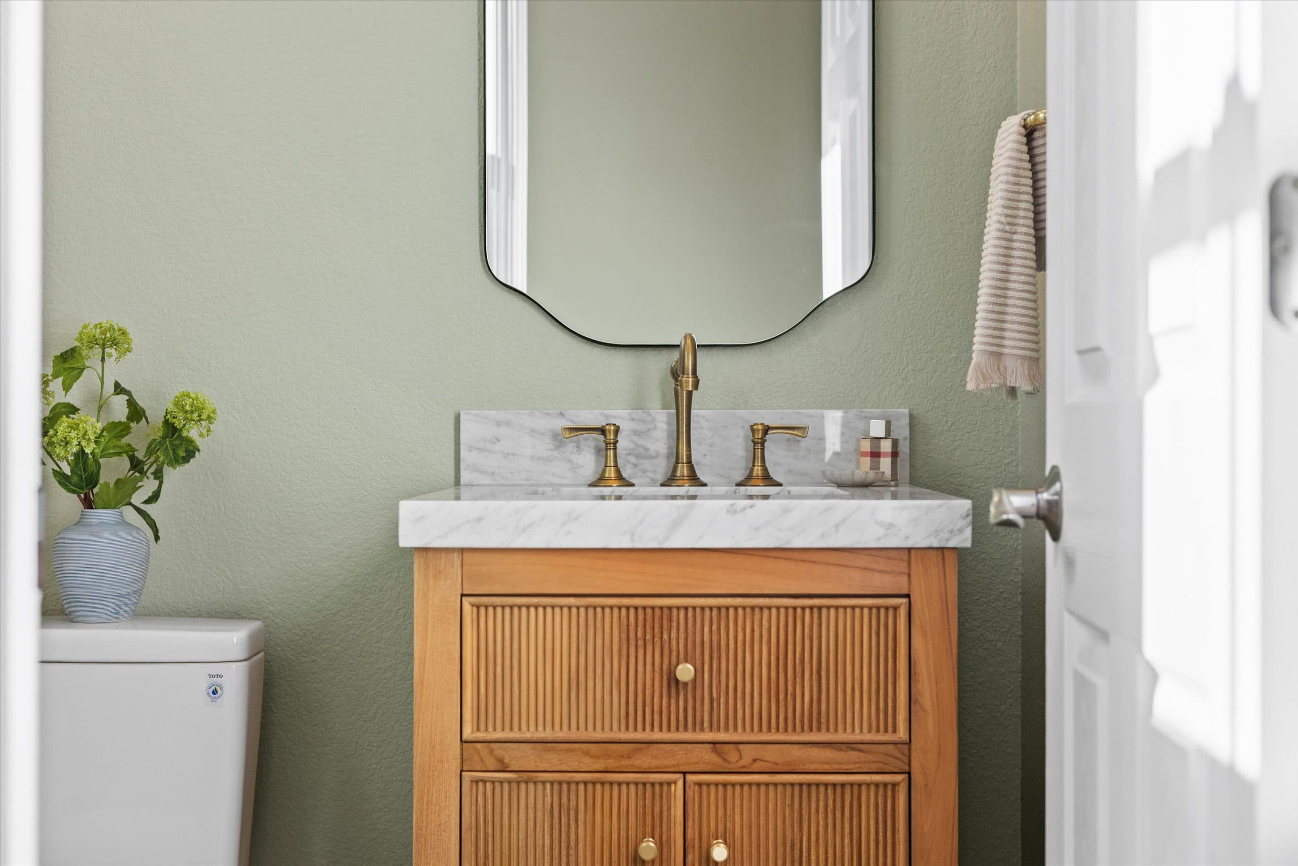 Close-up of powder bath vanity with natural wood cabinet, white marble countertop, brass faucet, arched mirror, sage green walls, and decorative vase with greenery