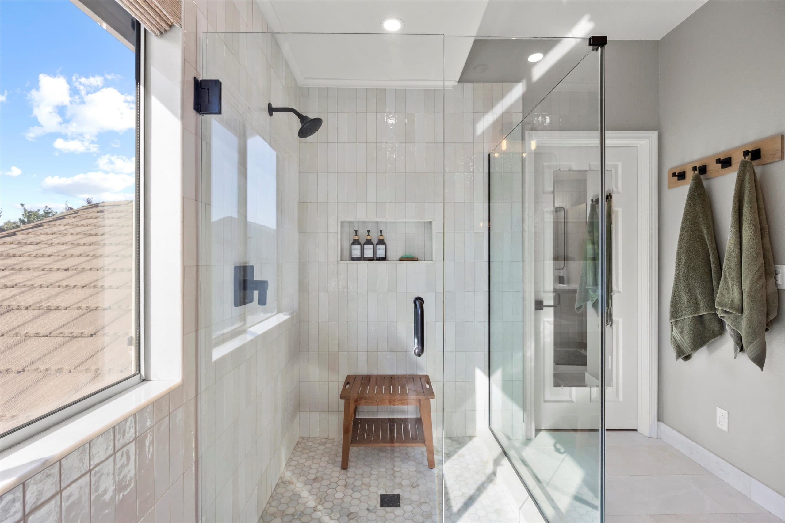 Exterior view of master bath shower enclosure with black-framed glass door, white subway tile, built-in niche, teak shower bench, hexagonal floor tile, and matte black rain shower head