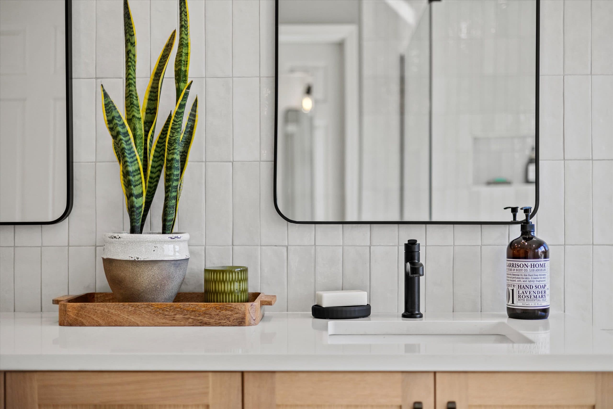 Close-up vanity vignette with snake plant, wooden tray, matte black faucet, amber hand soap dispenser, black-framed mirror edge, and white subway tile backsplash