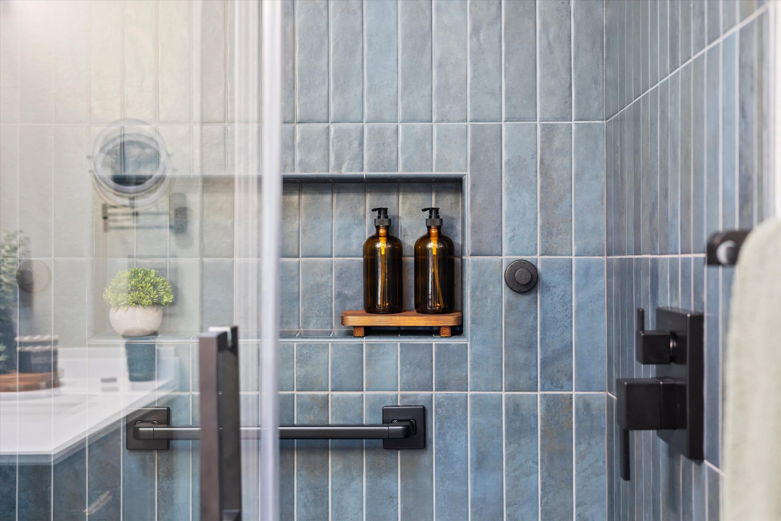 Close-up of shower niche with wooden shelf and soap dispensers set into blue subway tile wall with matte black fixtures