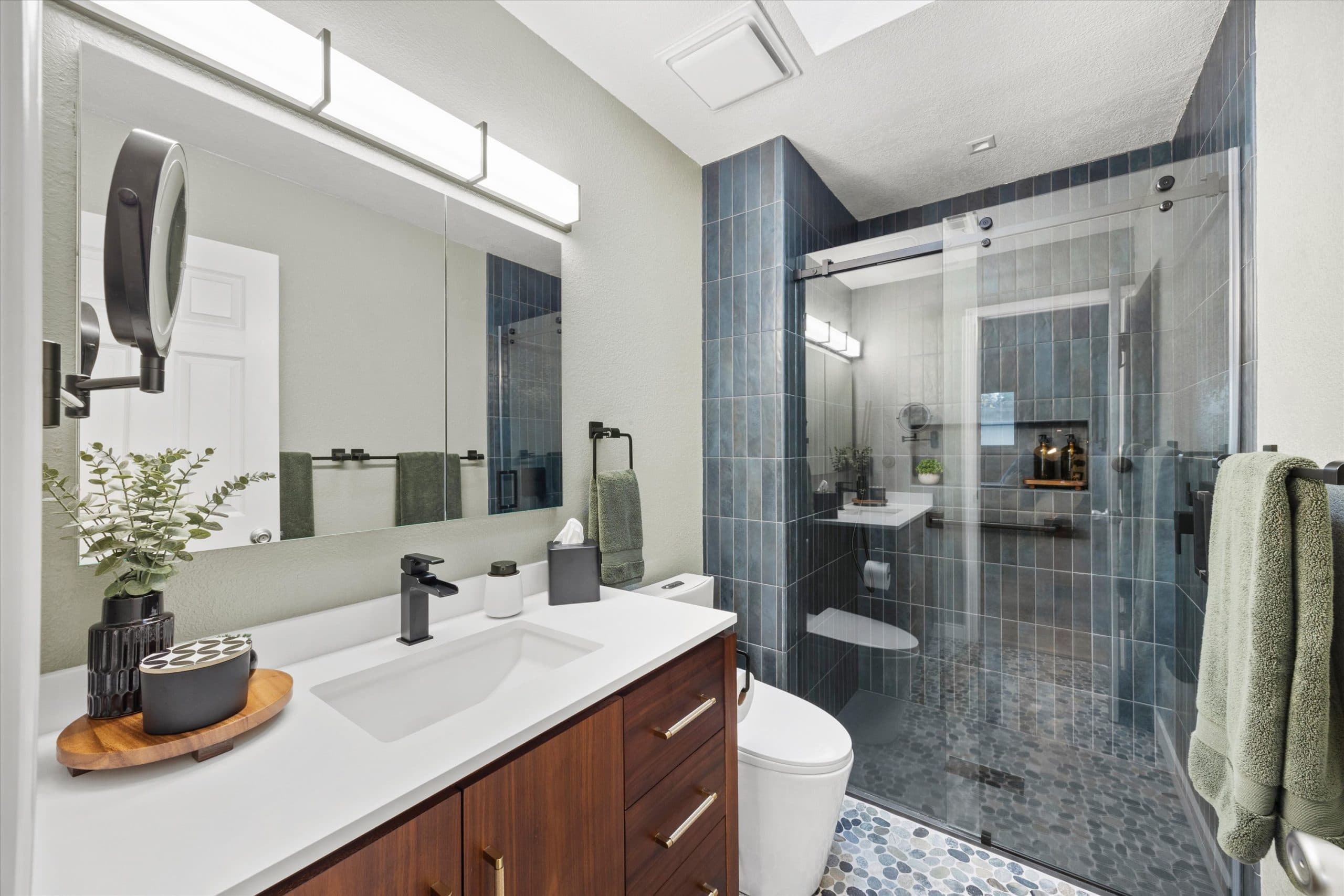 Bathroom remodel showing vanity area with round mirror and matte black hardware alongside glass-enclosed shower with blue tile