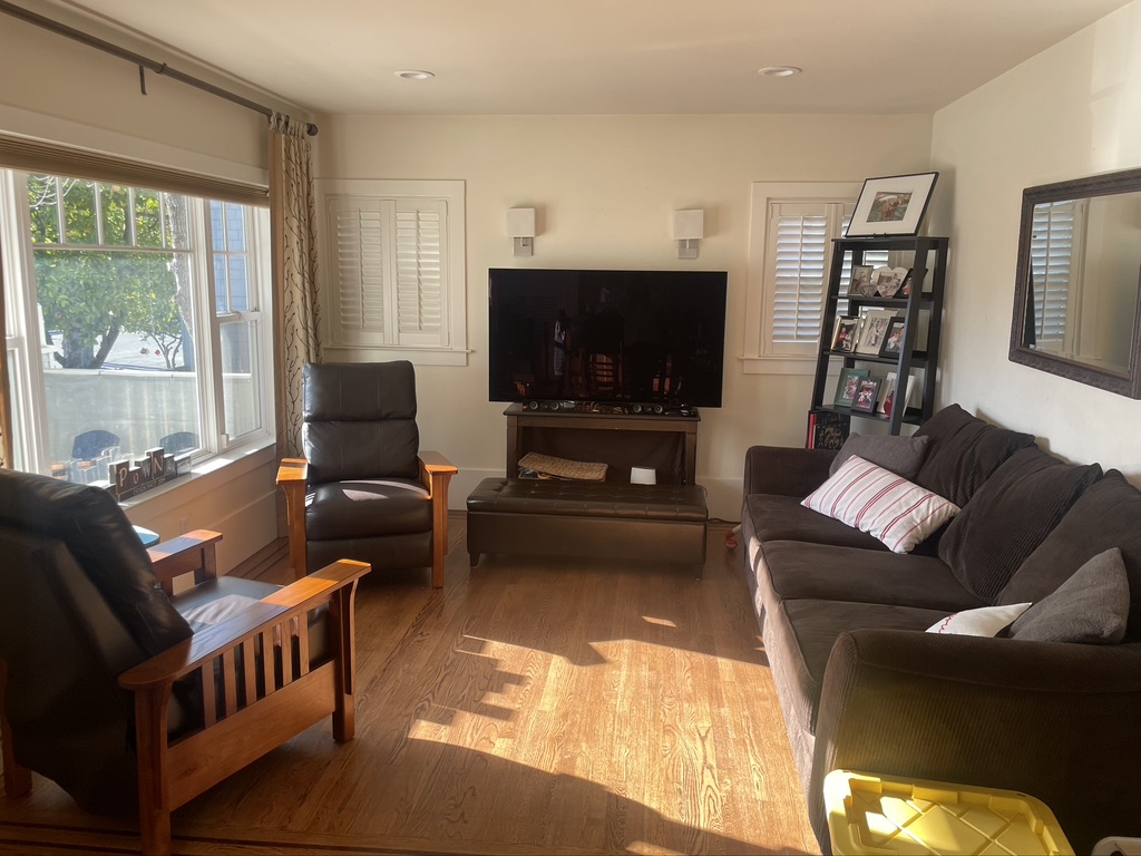 Before renovation, a living room with a couch, chair, and TV, highlighting the existing fireplace area.